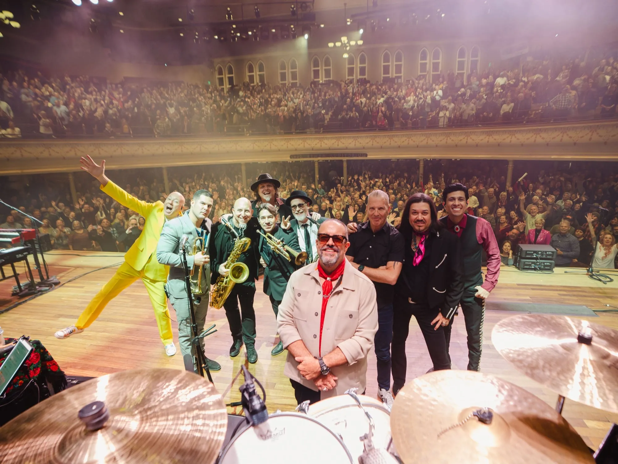 Full band and crew celebrating on stage, packed multi-tiered concert hall audience