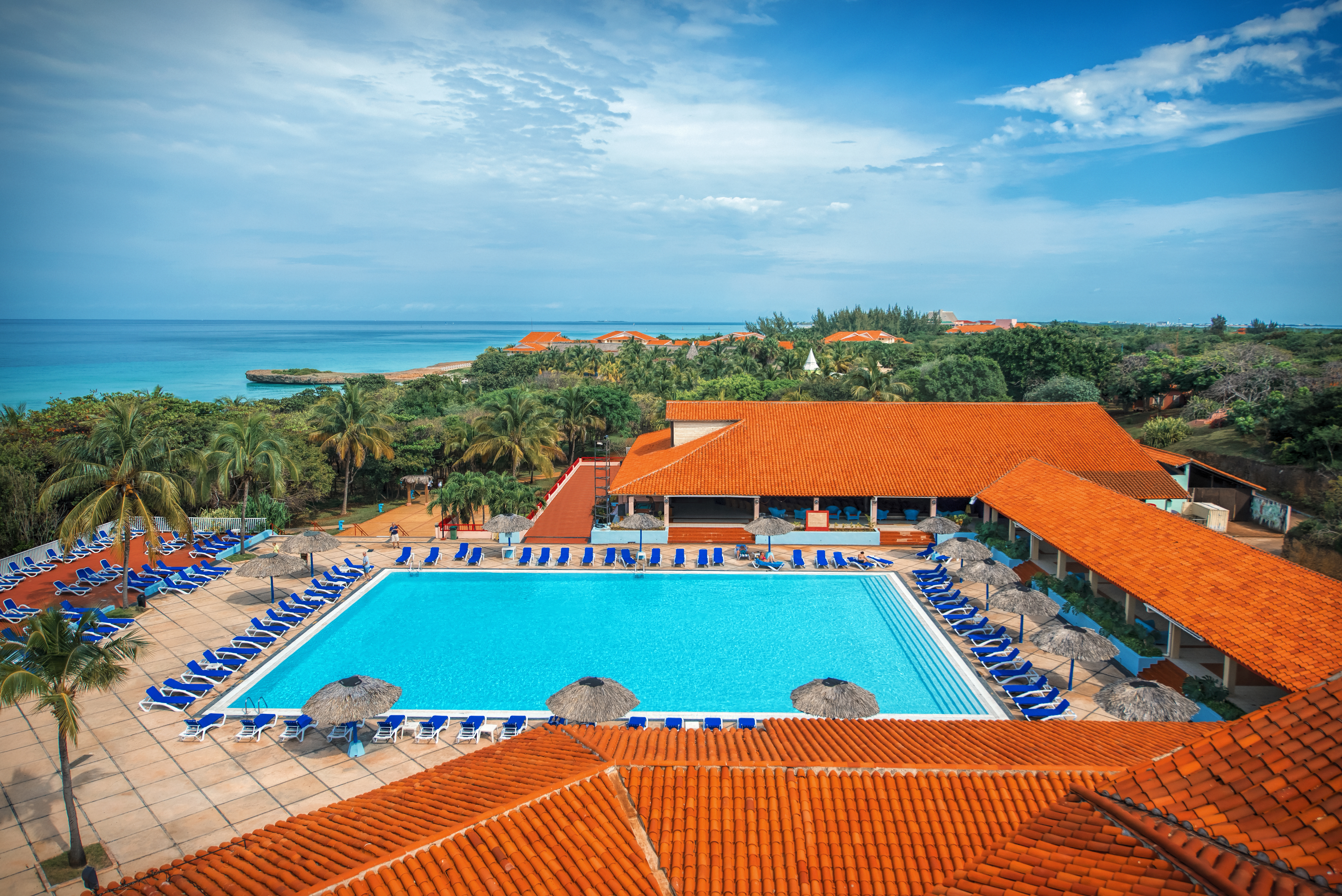 Aerial view of oceanfront resort property — large pool, terracotta rooftops, and turquoise water beyond